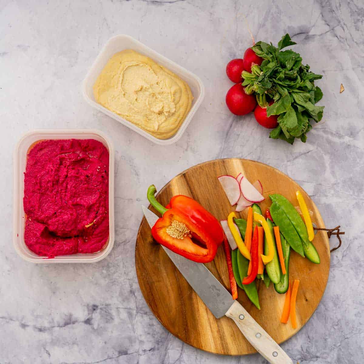 A wooden chopping board sitting on the bench with a knife and cut up vegetables sitting next to a container of hummus and beetroot hummus and radishes.