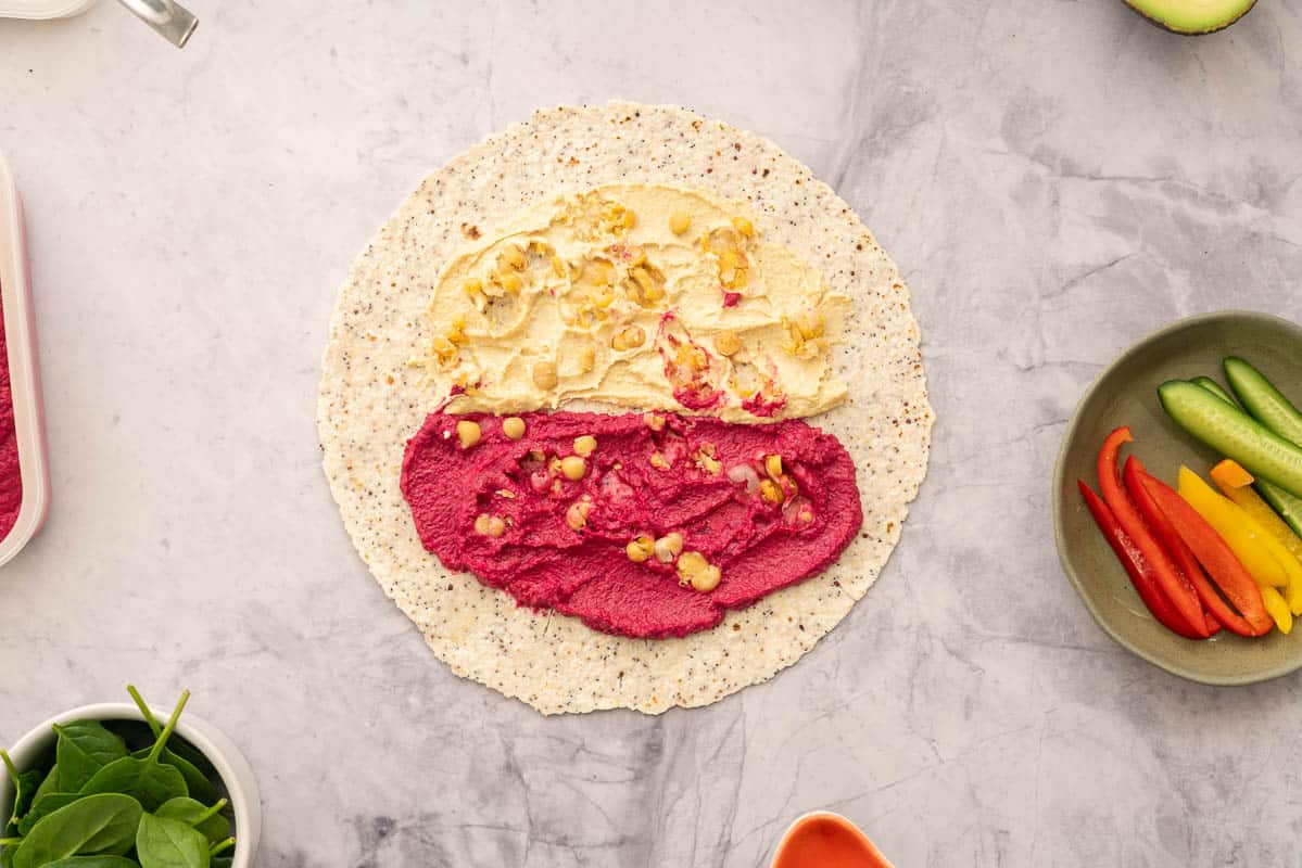 A round wholegrain wrap spread with beetroot hummus and regular hummus, topped with chickpeas, sitting next to individual ramekins of vegetables