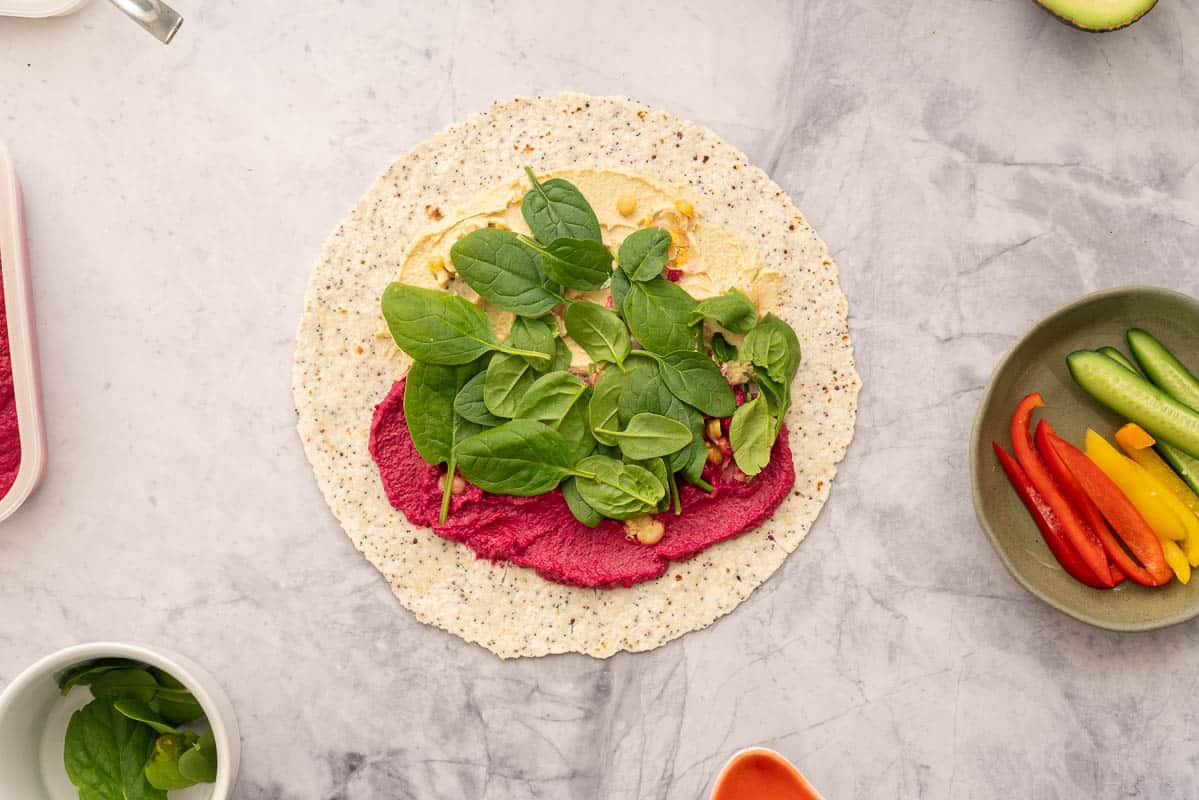 A round wholegrain wrap laid out on the bench spread with beetroot hummus and regular hummus, topped with chickpeas and spinach sitting next to individual ramekins of vegetables