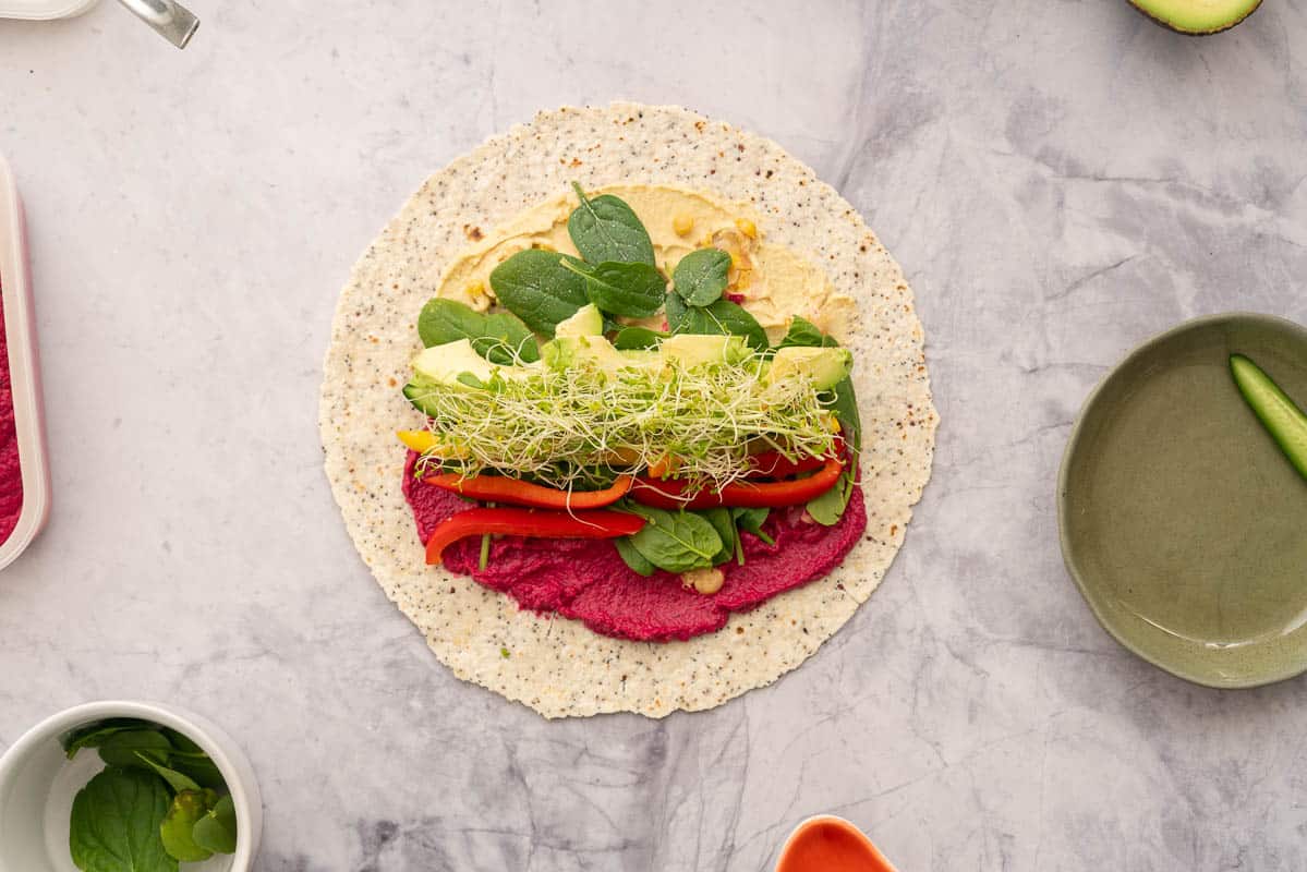 A round wholegrain wrap spread with beetroot hummus and regular hummus, topped with chickpeas, spinach, sliced vegetables and sprouts sitting next to individual ramekins of vegetables