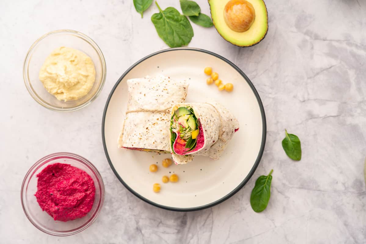 Halved filled wrap stuffed with beetroot hummus, cucumber, sprouts, spinach and capsicum sitting on a plate on a bench with an avocado cut open, hummus and beetroot hummus on bench top.
