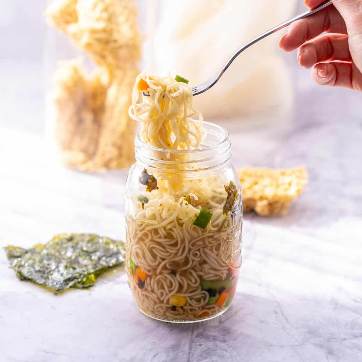 A glass jar filled with noodles and broth, a fork lifting the noodles up out of the jar, sheets of seaweed and a cake of dried noodles in the background. 