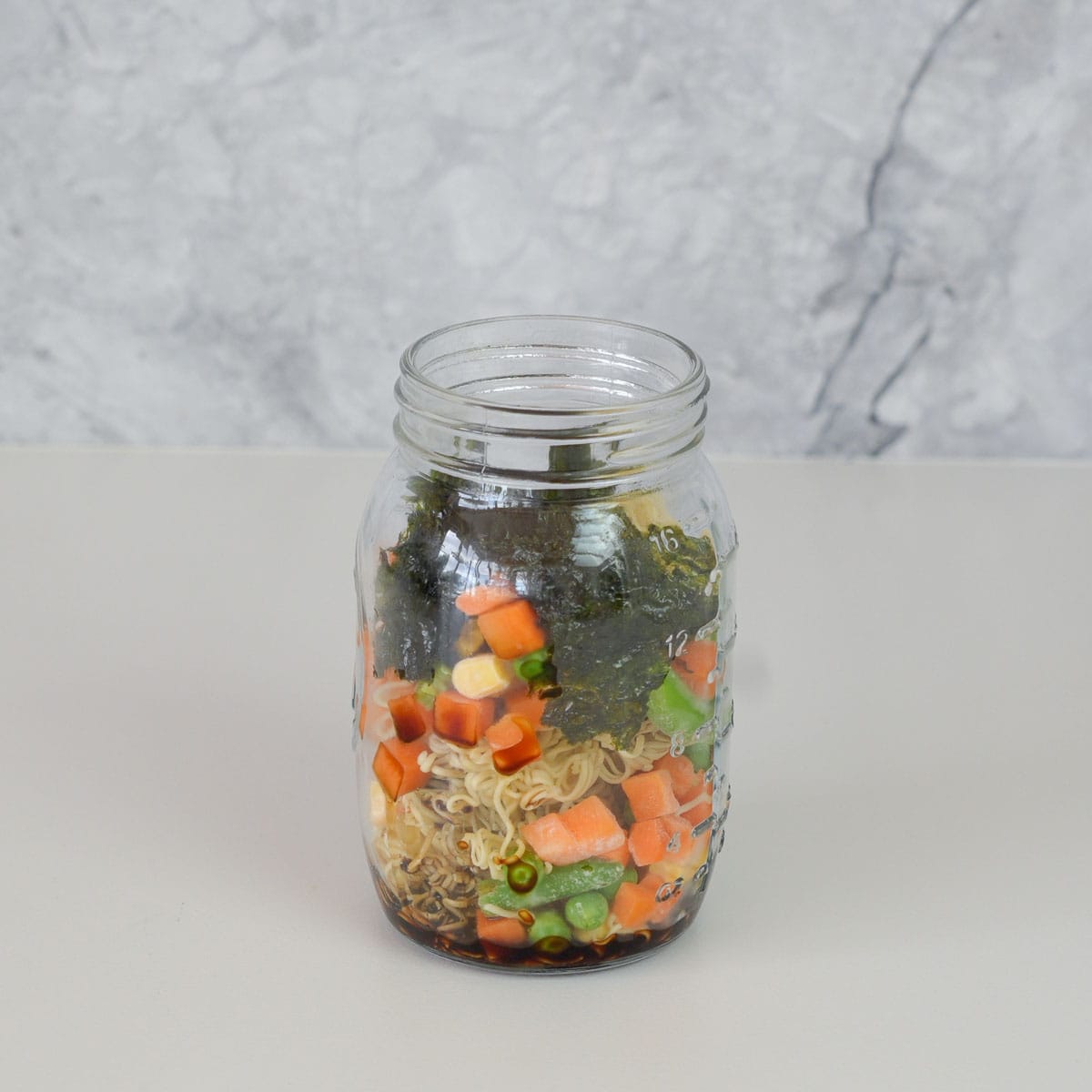 A mason jar on the bench with noodles, chopped carrot, corn kernels, beans and seaweed sheets teared up inside of it.
