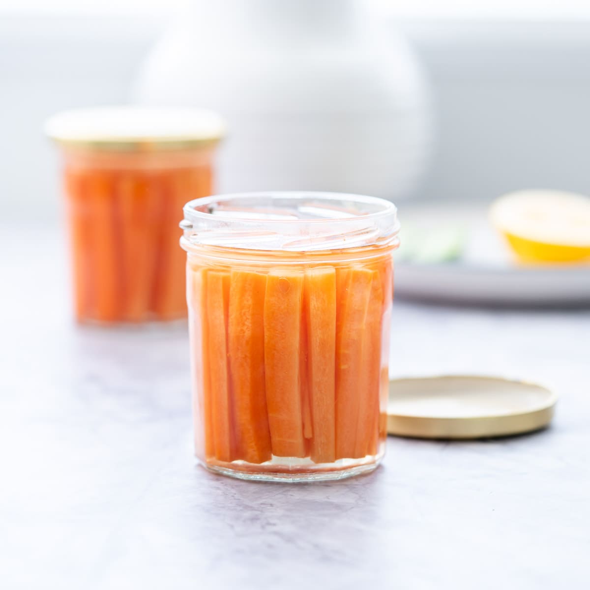 One jar with gold lid off filled with battened carrots covered in pickling juice sitting on a bench"