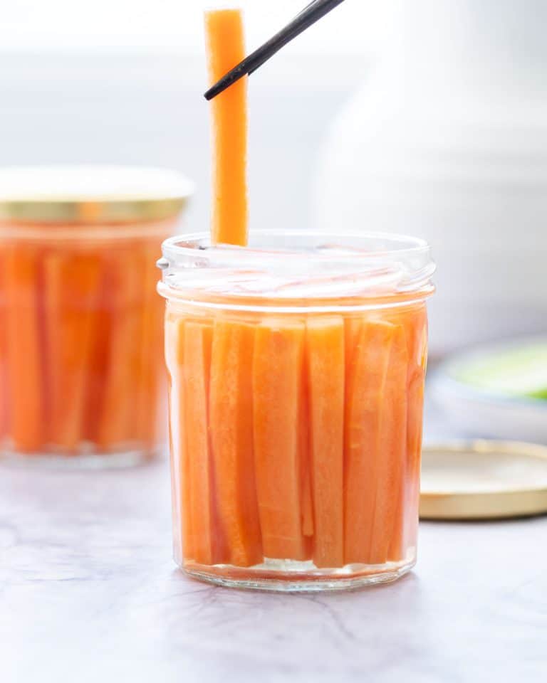 A carrot baton being lifted from a jar of pickled carrots with chop sticks.