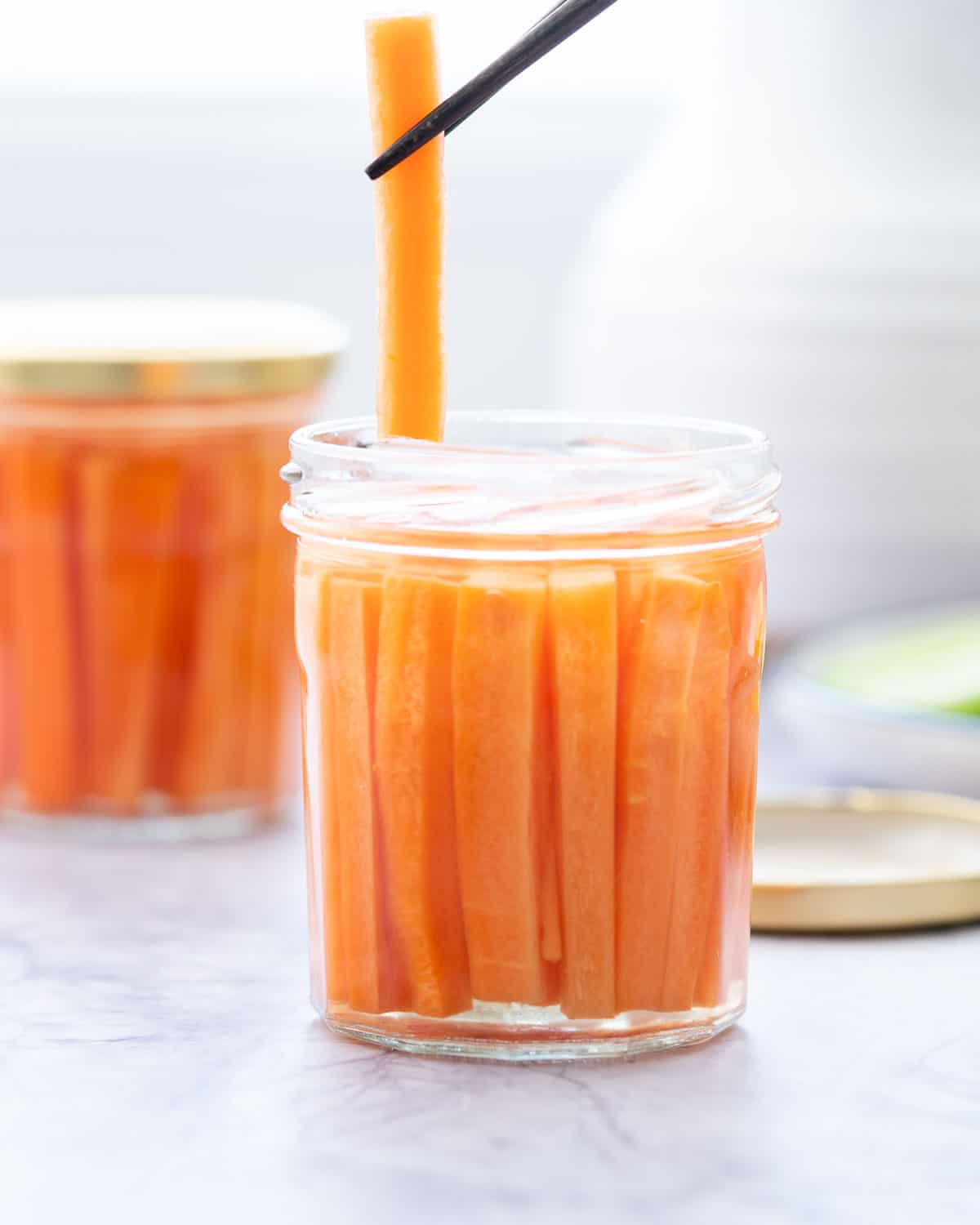Two glass jars filled with battened carrots covered in pickling juice sitting on a bench with one carrot batten being held above the jar with a chopstick