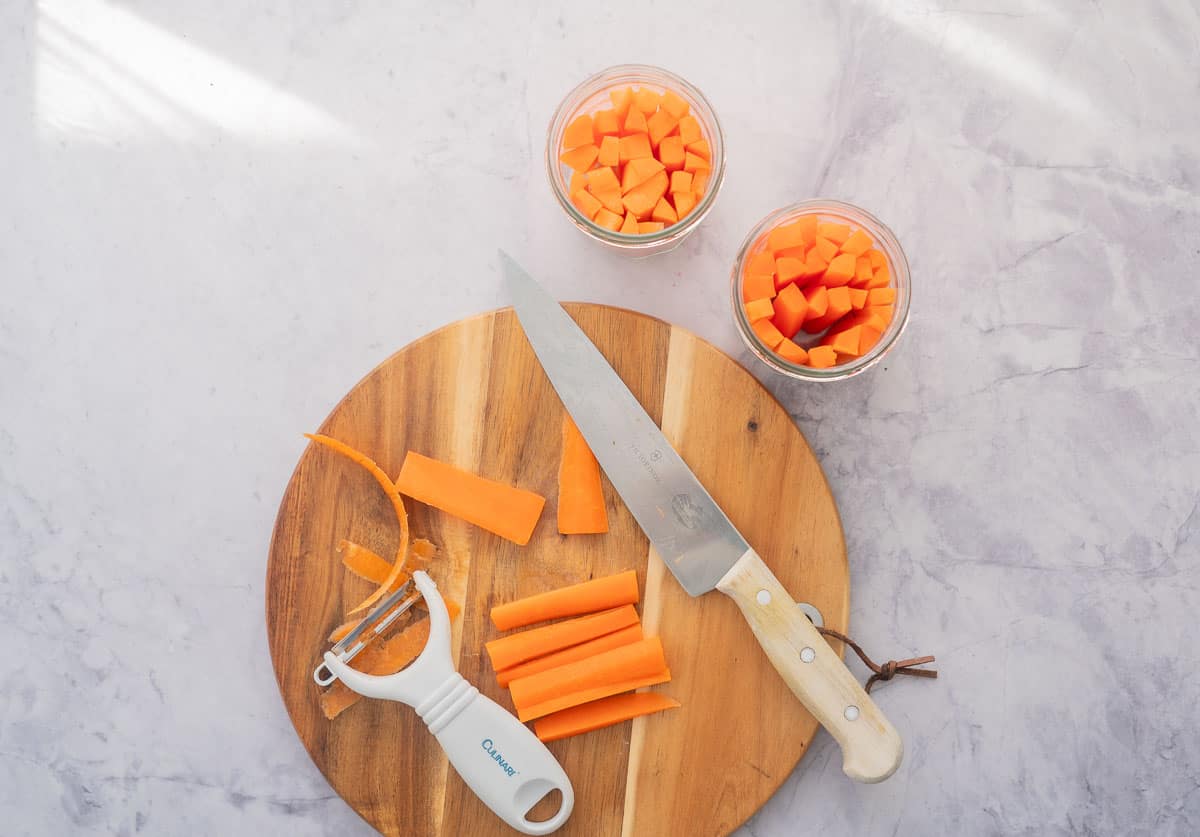 2 glass jars filled with battens of carrots, sitting next a chopping board with sliced carrots a knife and a peeler on it.