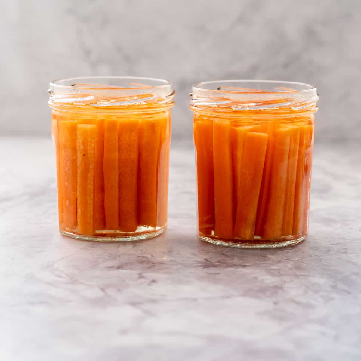 Two glass jars filled with battened carrots covered in pickling juice sitting on a bench