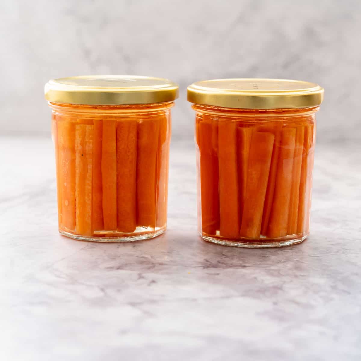 Two glass jars with gold lids screwed on filled with battened carrots covered in pickling juice sitting on a bench