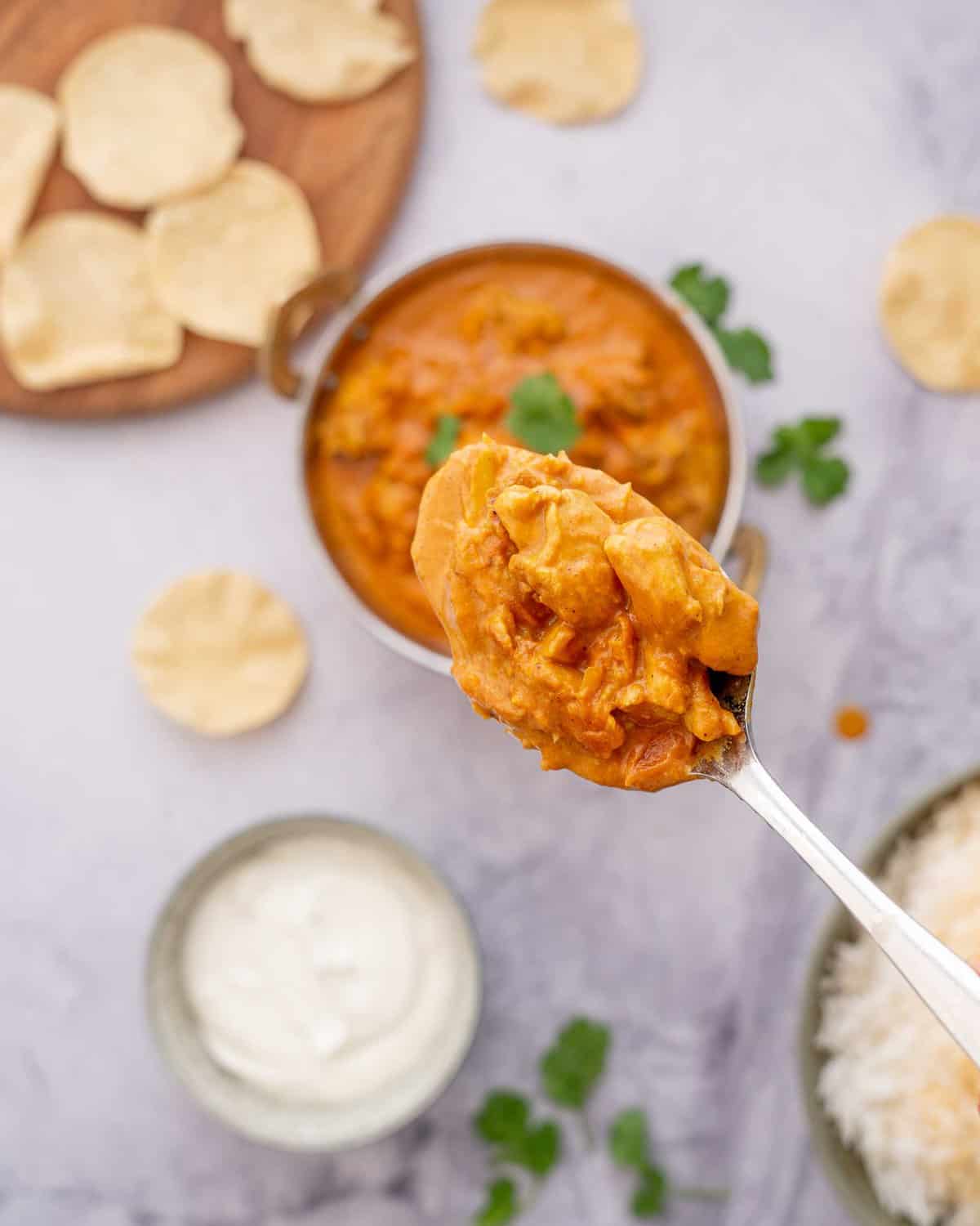A spoon full of butter chicken raised above a big bowl of rice and popadoms and herbs