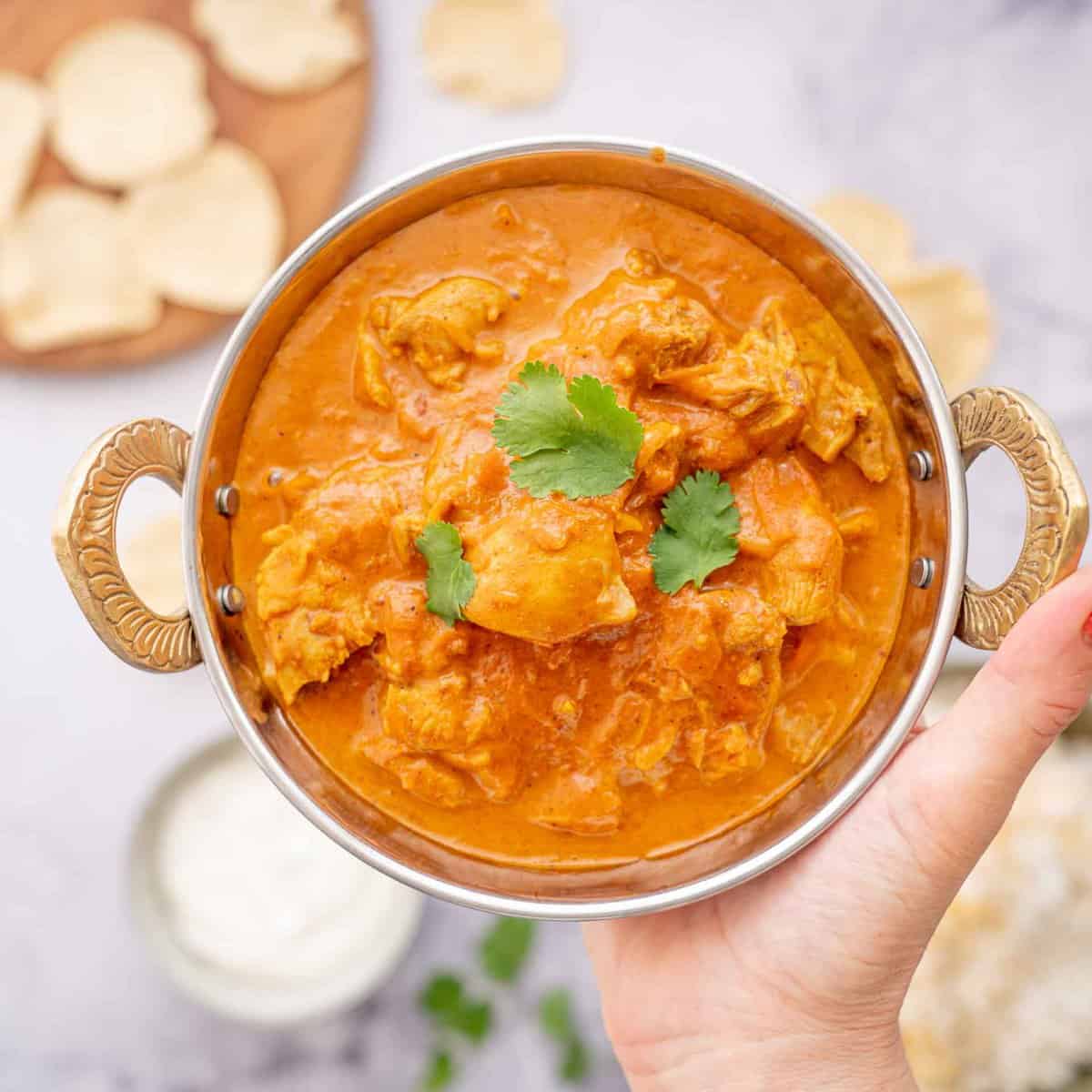 A hand holding up a bowl of butter chicken above a bench with bowls of rice and yoghurt 