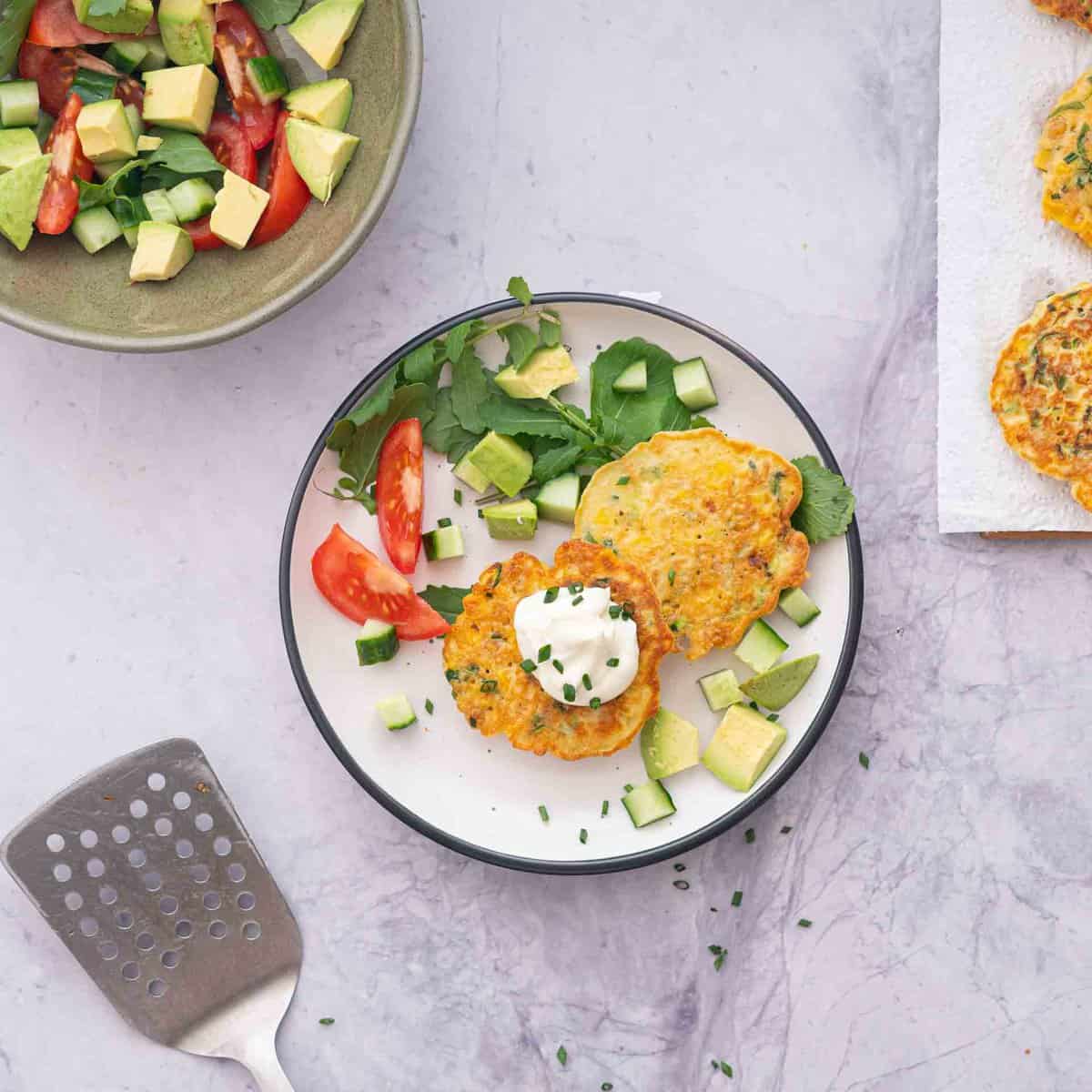 Two Chickpea fritters served on dinner plate with sliced tomato, chopped avocado and garnish.