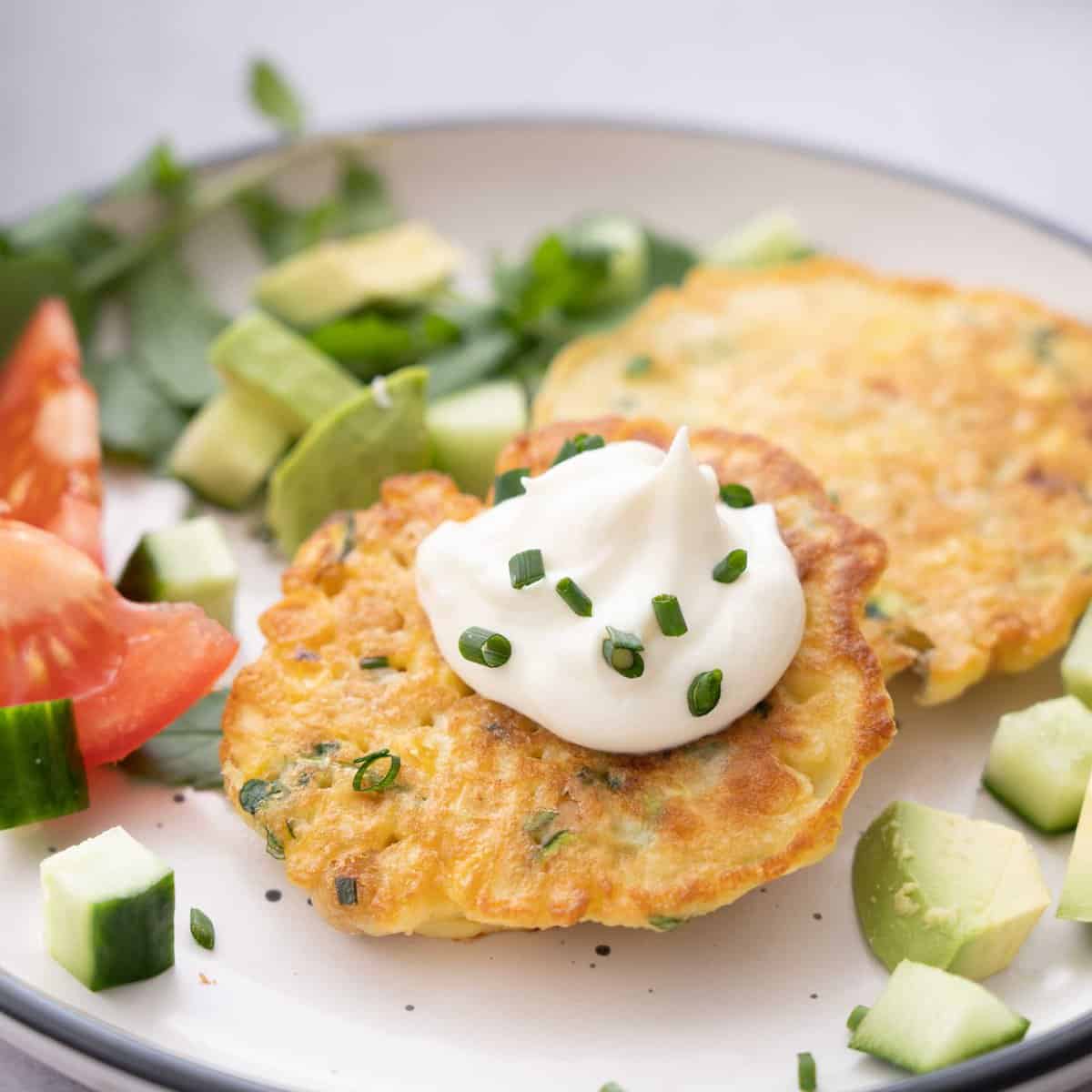 Two Chickpea fritters served on dinner plate with sliced tomato, chopped avocado and garnish.