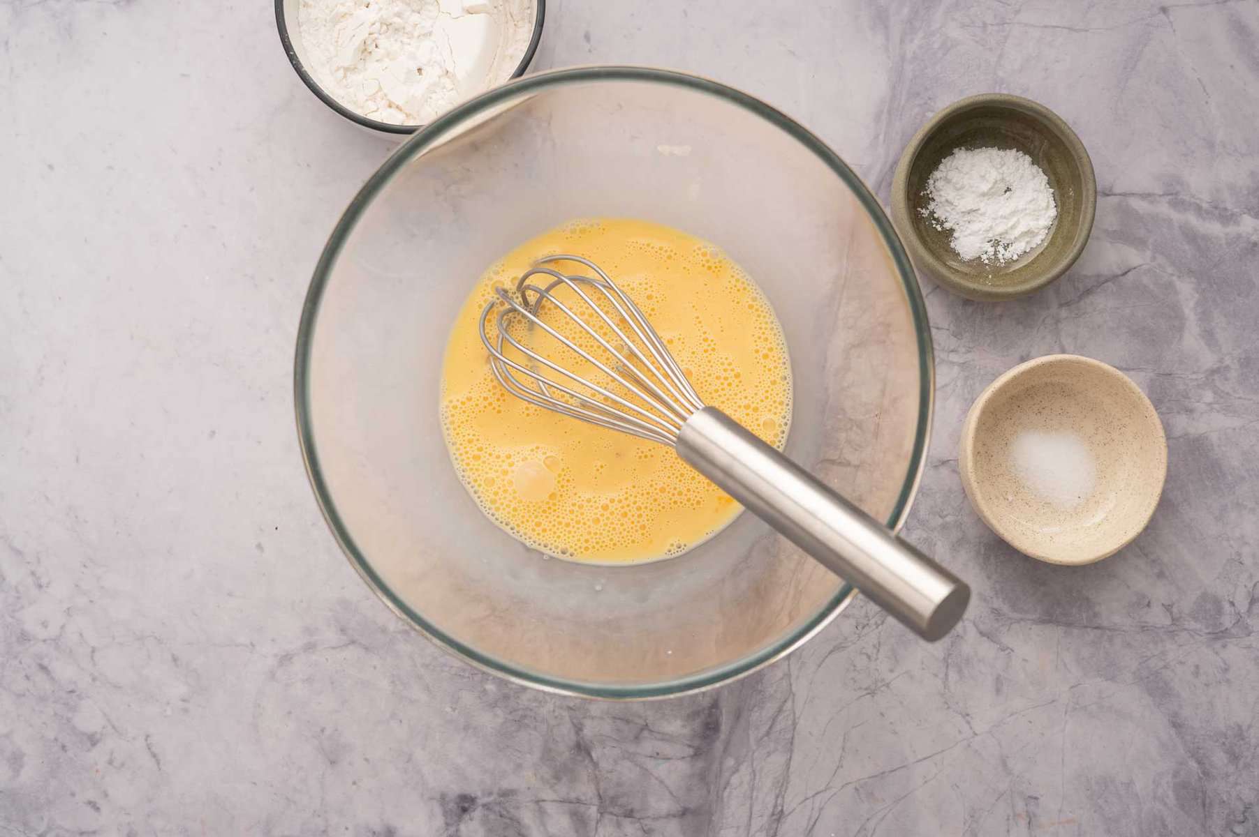 One large mixing bowl with an egg whisk and beaten eggs and 3 small ramekins individually filled with dry baking ingredients