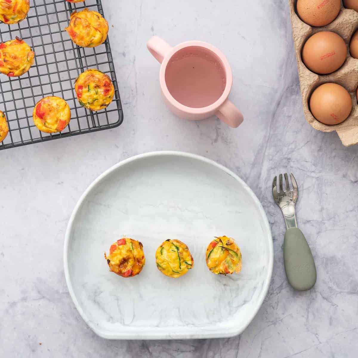 Three mini egg muffins on a silicone baby plate with a fork next to a baby sippy cup.