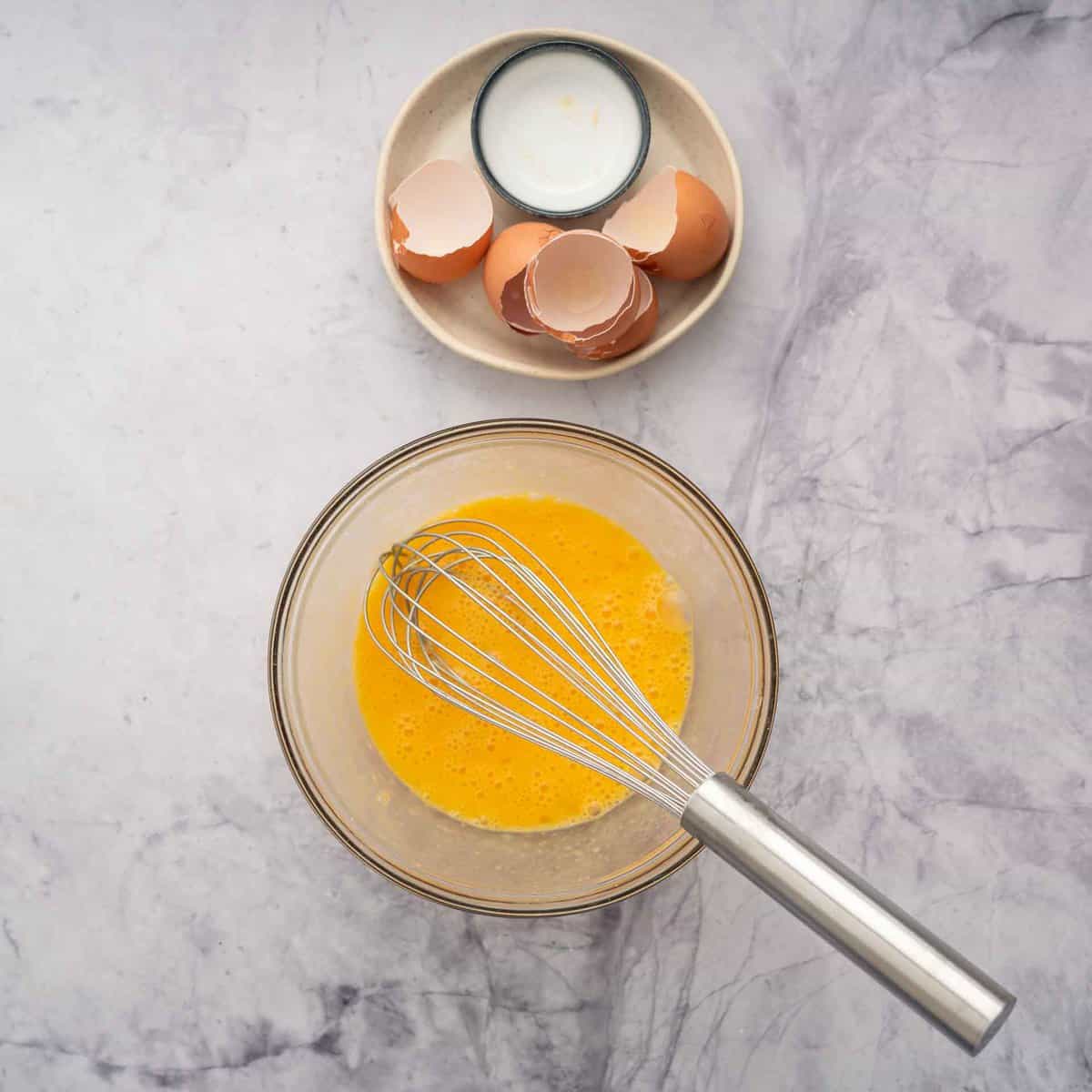 Whisked eggs in a glass mixing bowl with empty shells in seperate bowl.