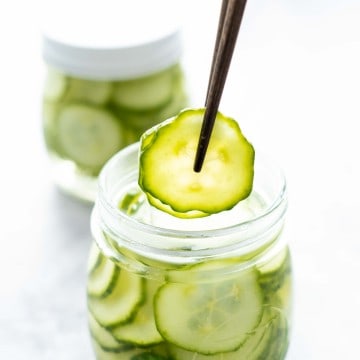Two slices of cucumber being held with chopsticks above a jar of pickled cucumbers.