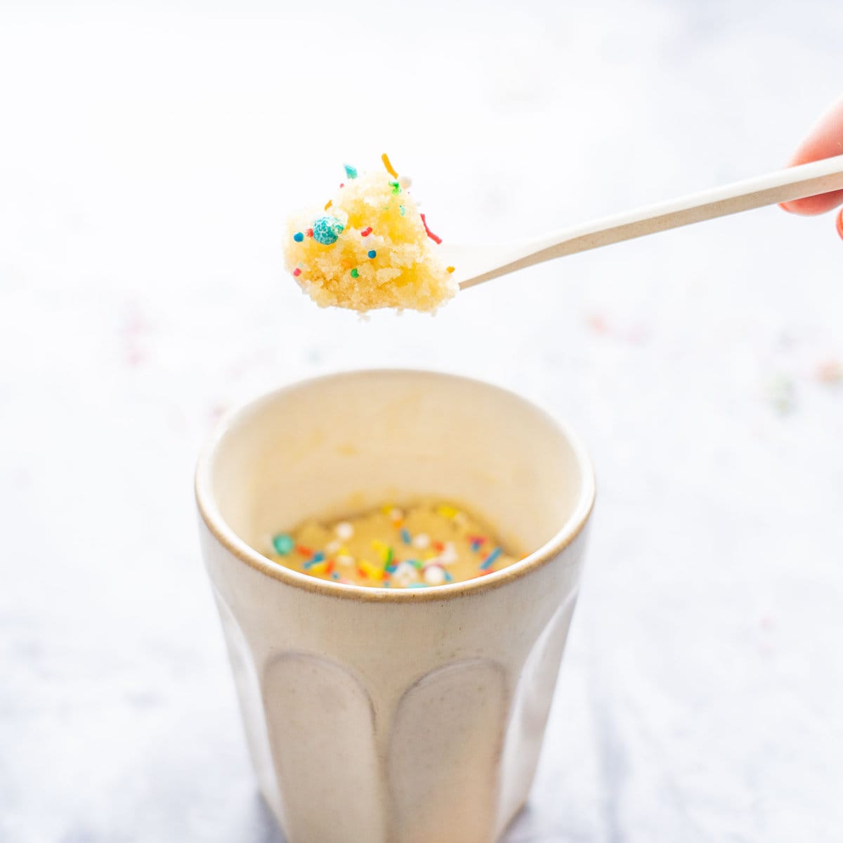 One vanilla Mug Cake sitting on the bench with sprinkles on it with a spoon raised above the mug with a mouthful of cake on it