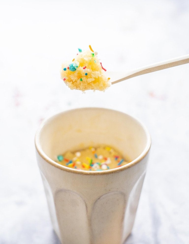 A spoon of vanilla cake decorated with sprinkles being held above a coffee mug