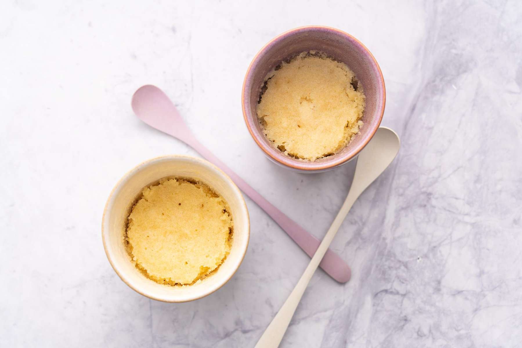 Two mugs next to each other showing the Vanilla Mug Cake cooked with two spoons laid out on the bench