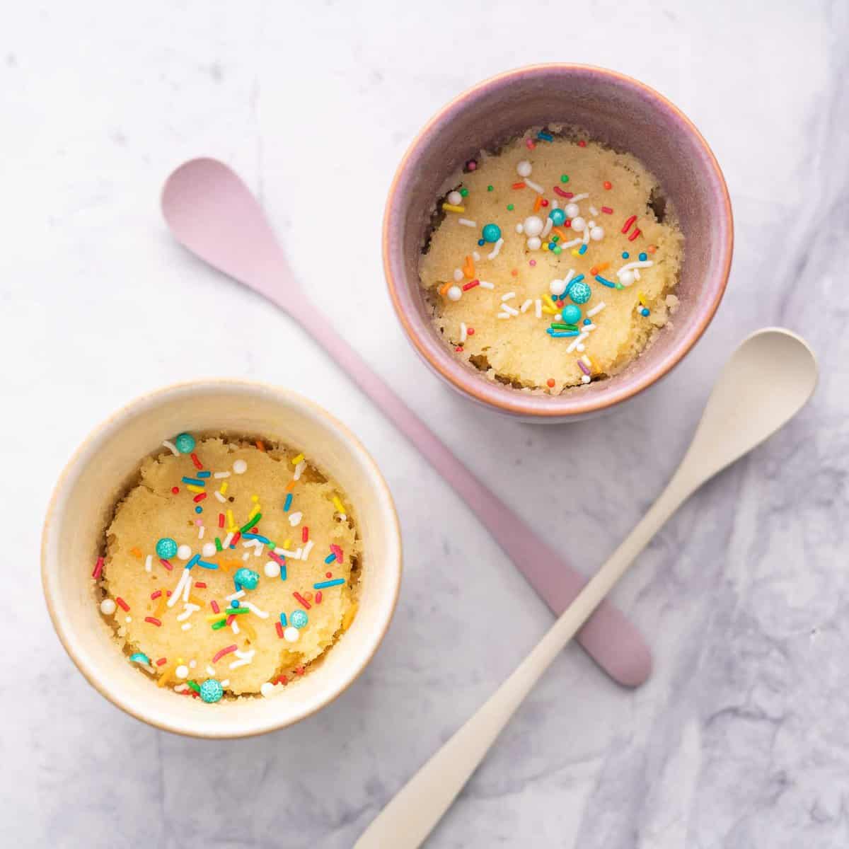 Two mugs next to each other showing the Vanilla Mug Cake cooked with scattered coloured sprinkles on top with two spoons laid out on the bench