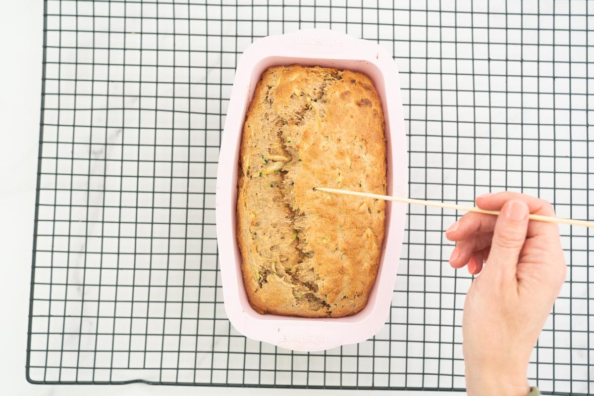A banana bread in a pink loaf tin being pierced with a bamboo skewer.