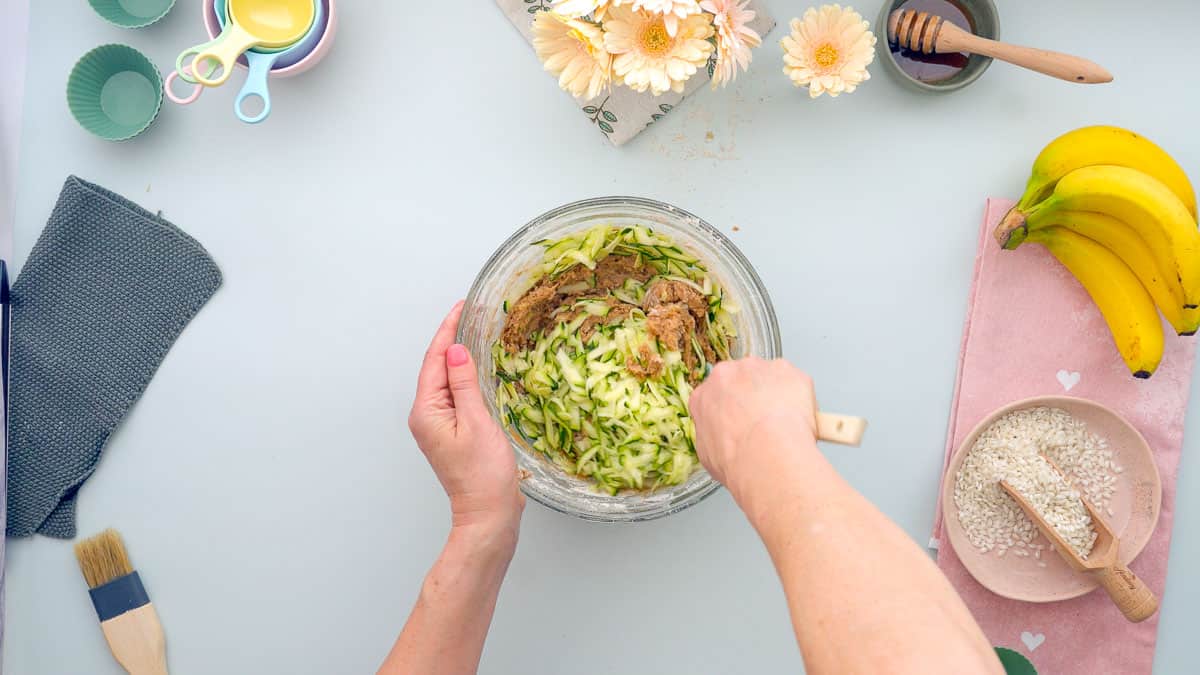 Grated zucchini being mixed through banana bread batter with a woodden spoon.
