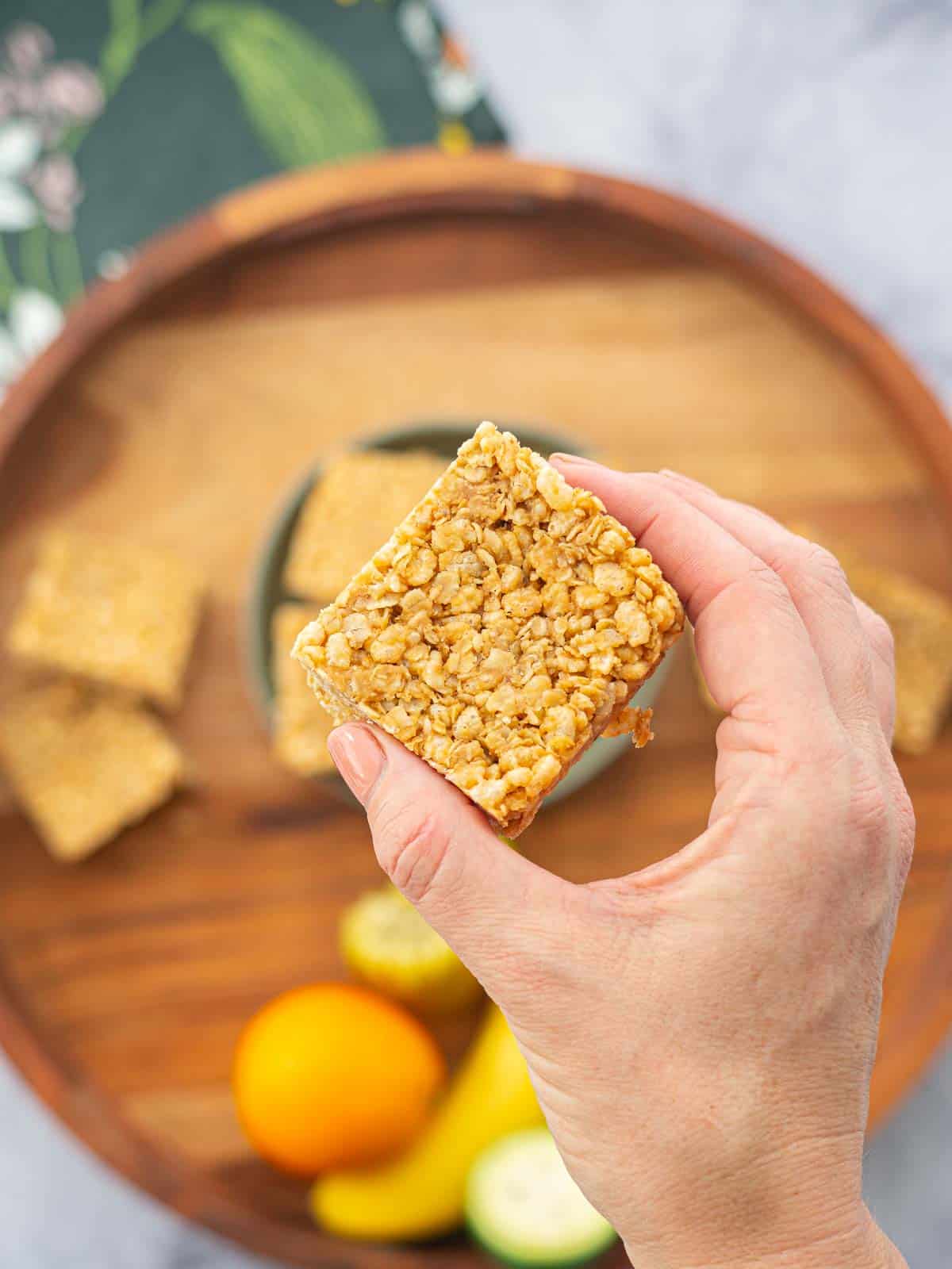 A piece of peanut butter slice being held above a wooden tray with fruit and more slice.