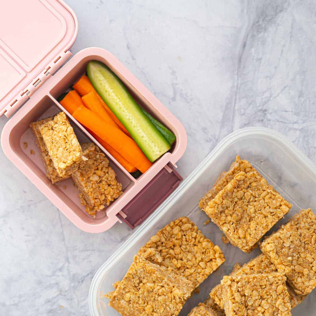A container of rice krispie treats next to a small pink bento box packed with rice krispie treats and vegetable sticks.
