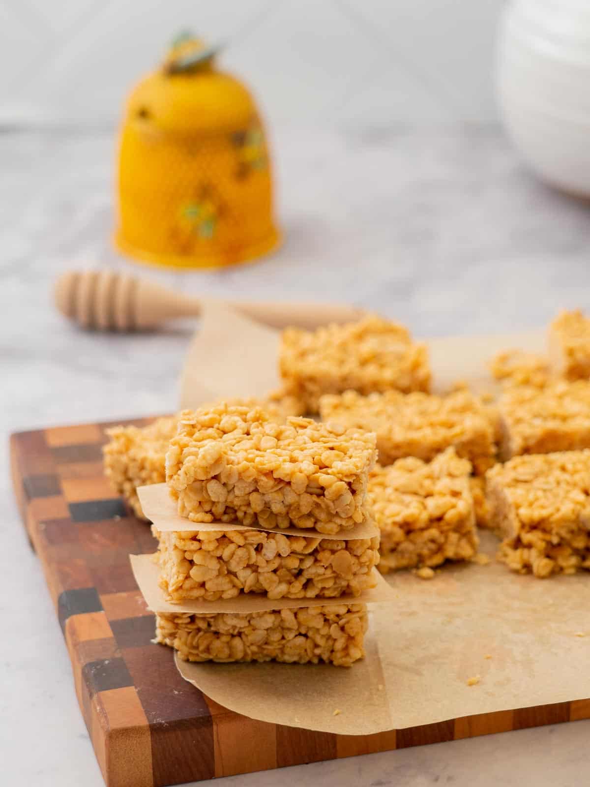 A tower of rice krispie treats on a wooden chopping board in front of a honey jar.