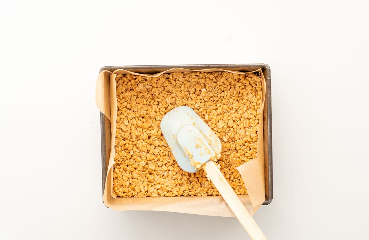 Rice krispie mix being pressed into the bottom of a lined baking tin with a spatula.