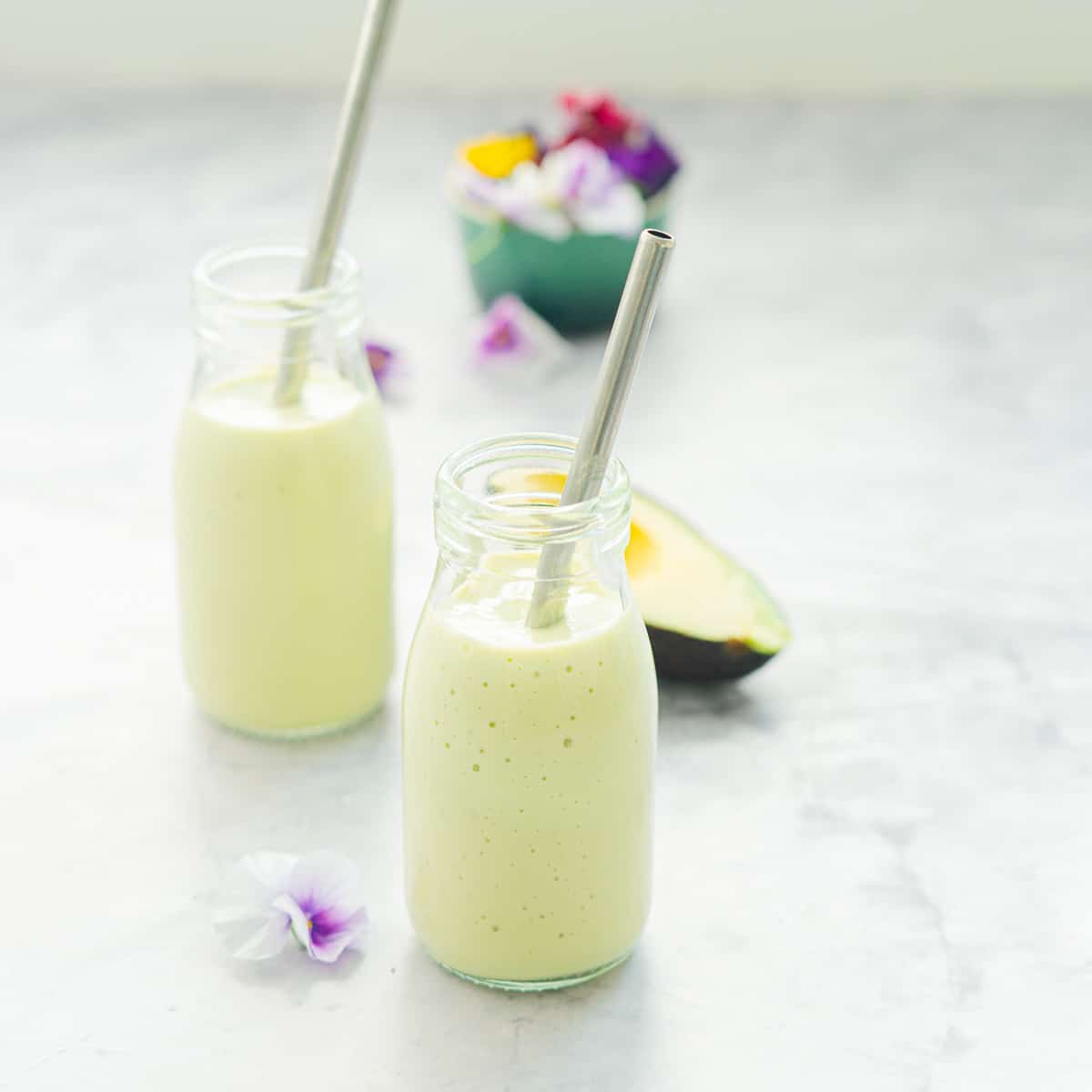 Two glass jars filled with Avocado Smoothie sitting on a bench with metal straws in each one as well as half an avocado and a small bowl of edible flowers in behind them.