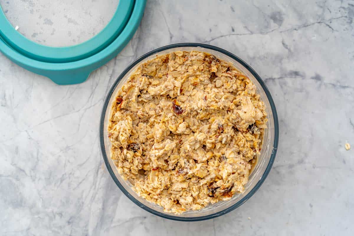 Large glass bowl filled with Bircher Muesli with a blue lid sitting on the bench next to it