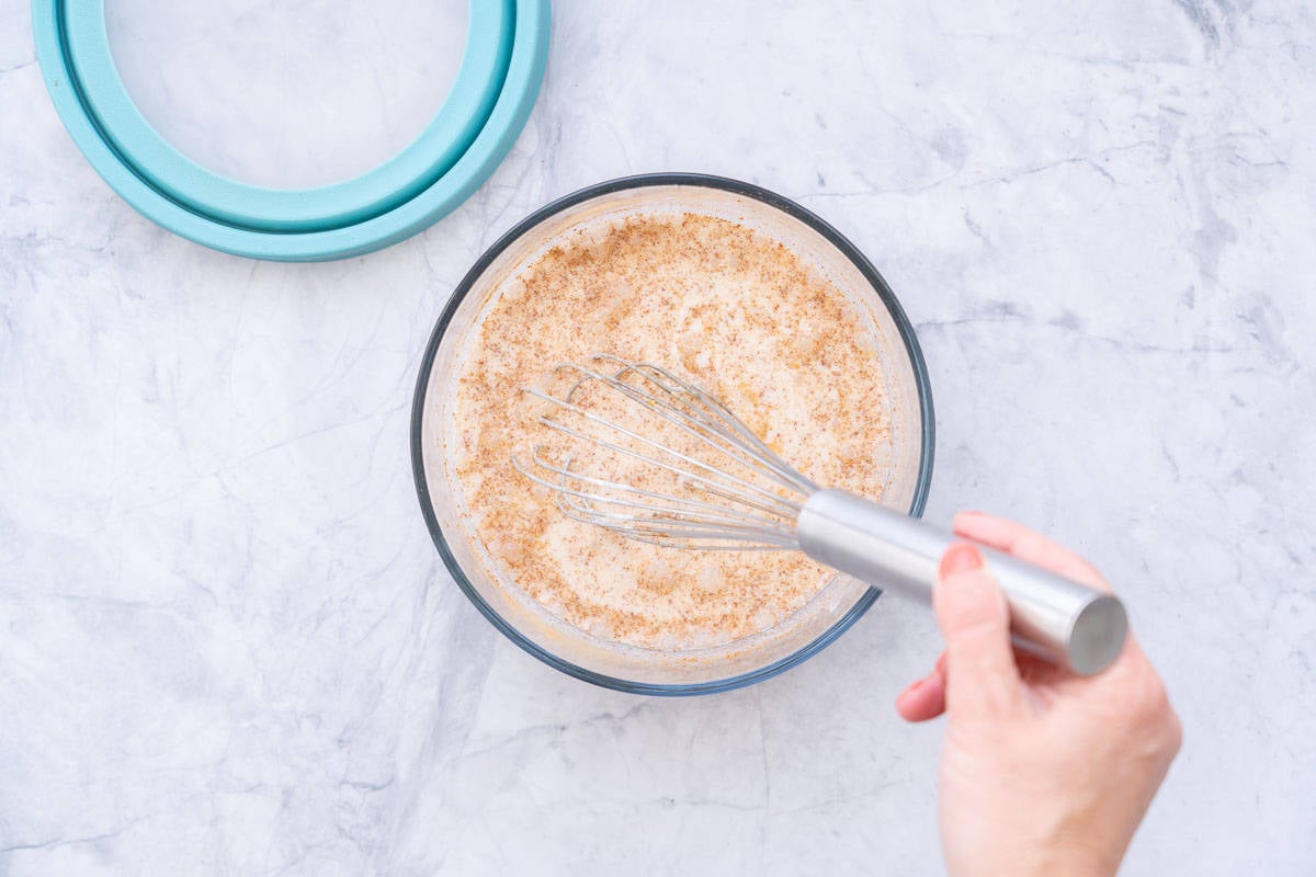 Whisked ingredients in a large glass bowl sitting on a bench with a blue lid next to it