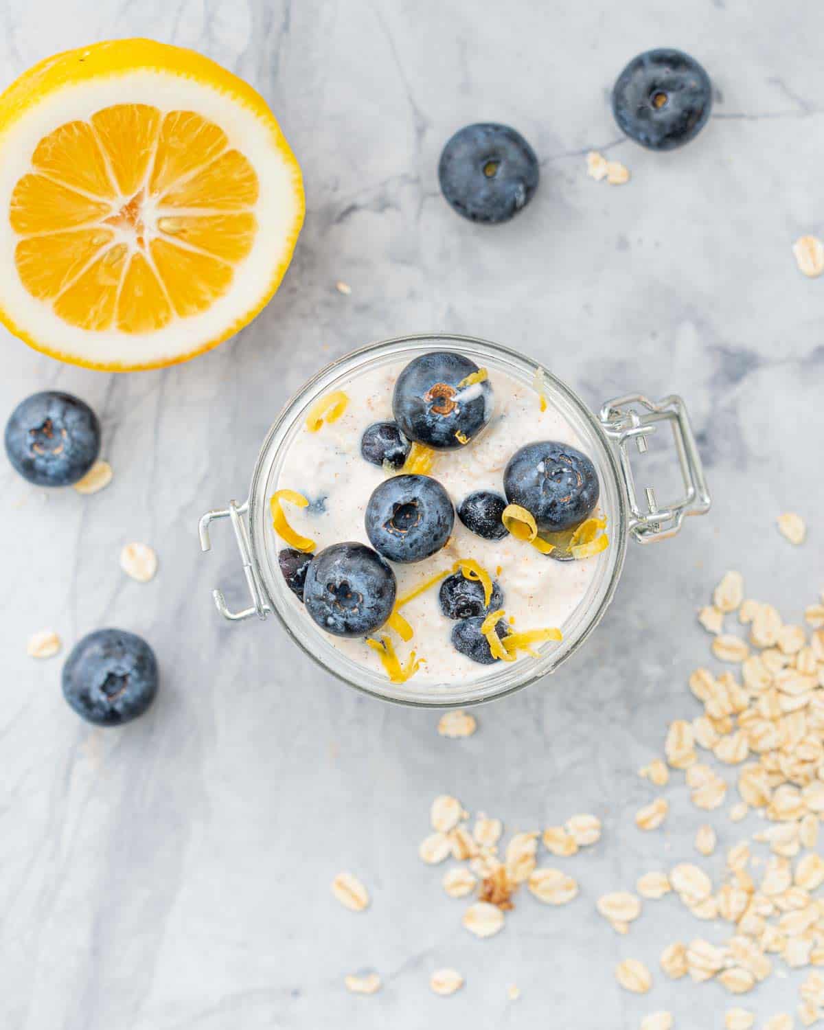 Looking down onto Blueberry Overnight oats in jar with fresh whole blueberries on top and grated lemon zest. Half a lemon and sprinkled oats and blueberries on bench top.