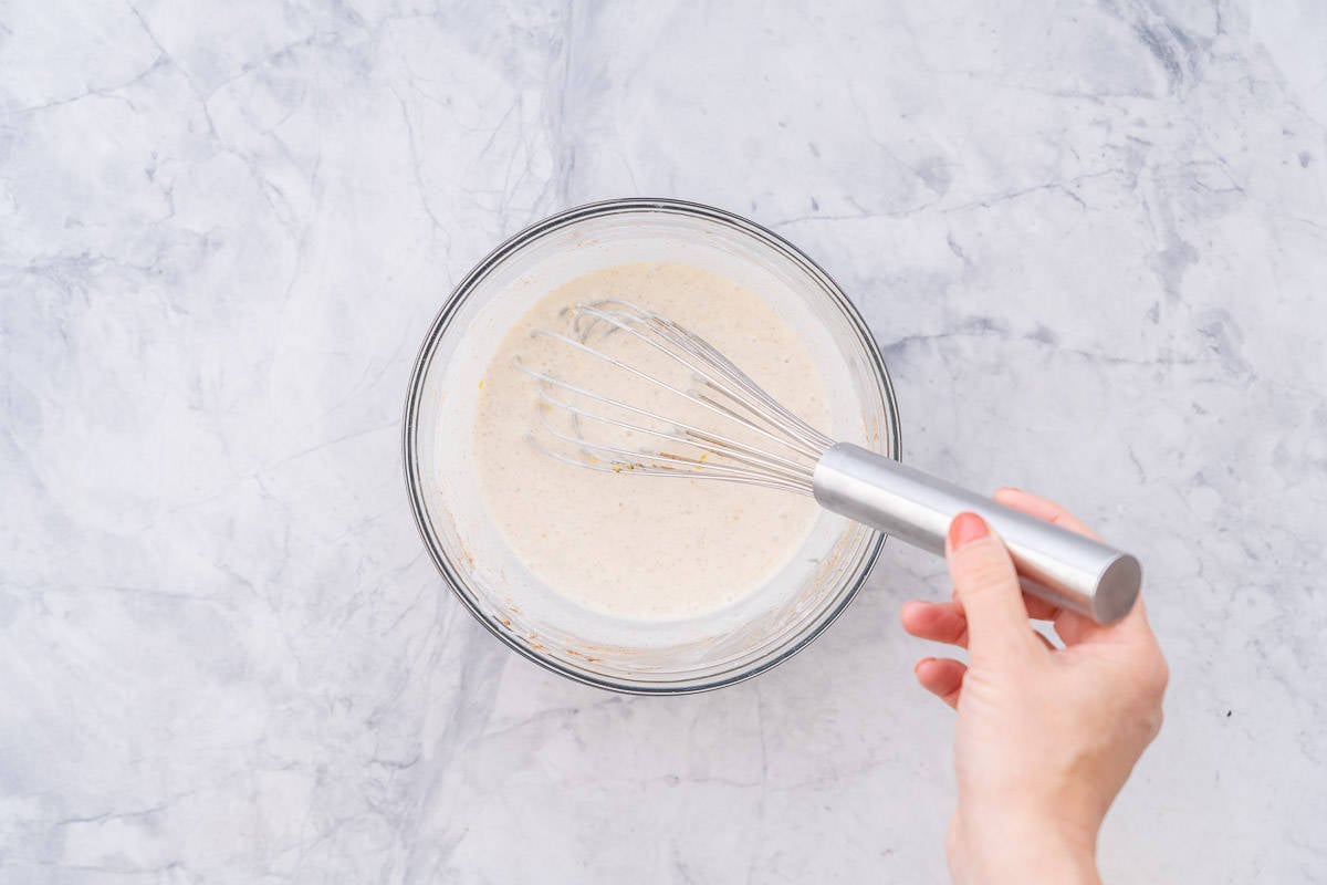 Whisked yoghurt, milk, maple syrup, cinnamon lemon juice and zest in a bowl on a bench with a hand holding a whisk in it.