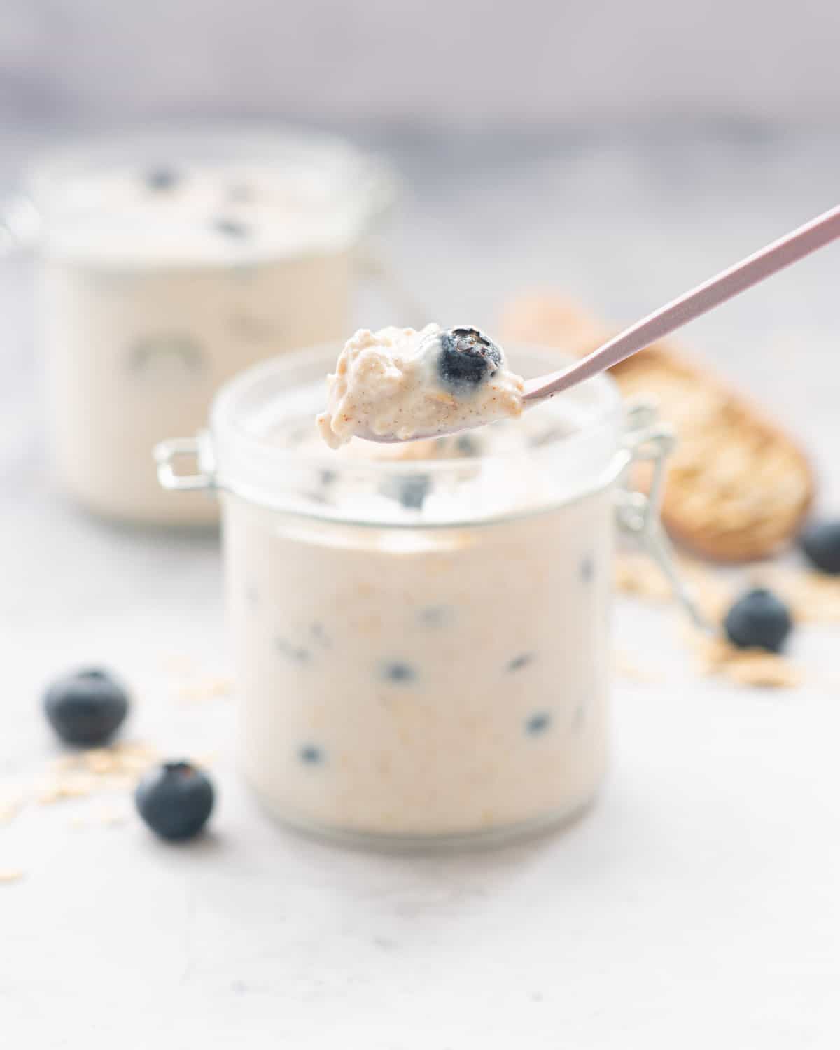 A spoonful of overnight oats held above two jars filled with Blueberry overnight oats sitting on a bench next to scattered blueberries and rolled oats.