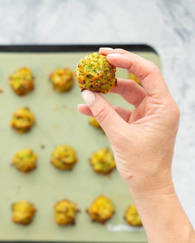 Cooked golden and crisp broccoli bites on a lined baking tray sitting on a bench with a hand holding one above them.