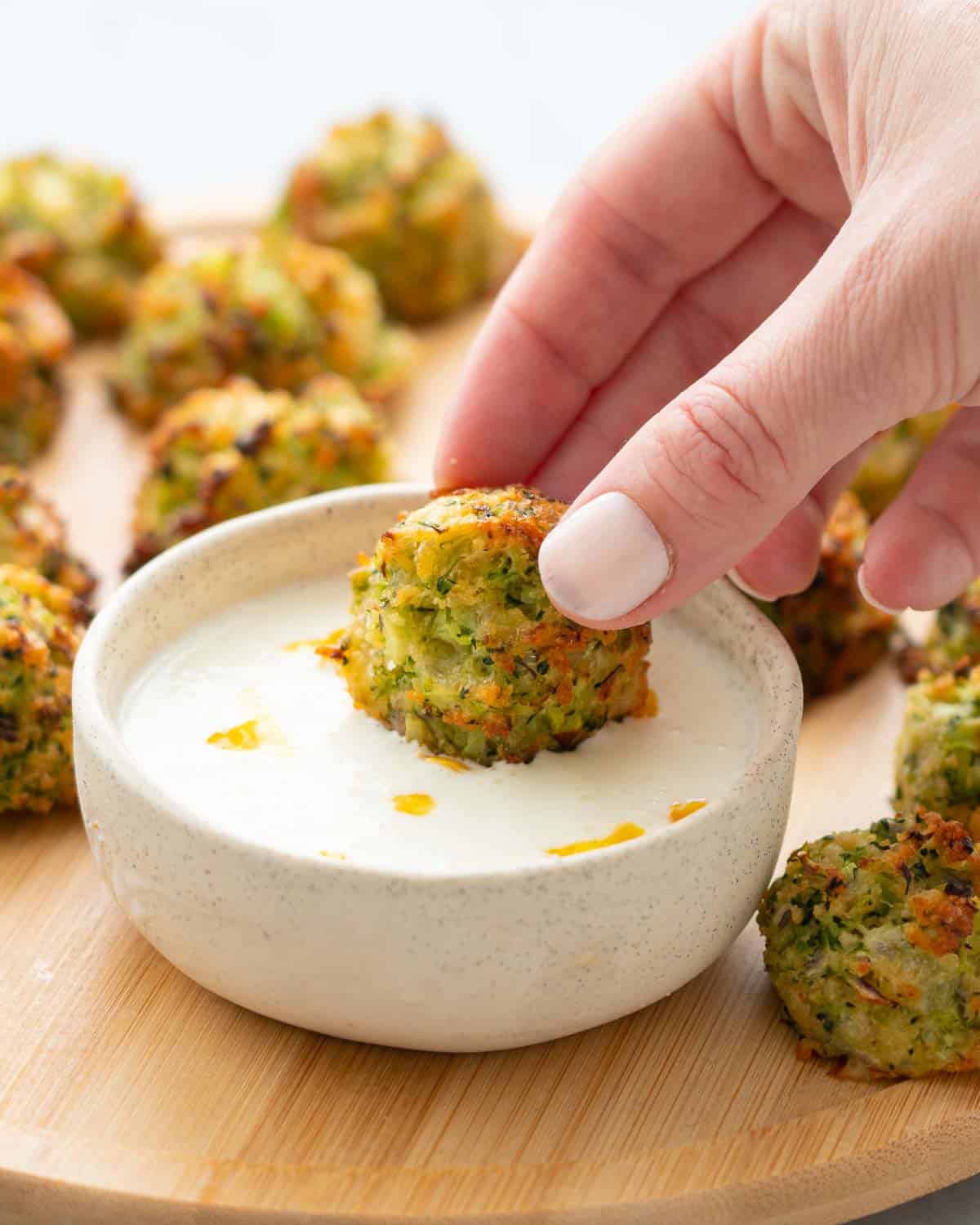 A hand dunking a broccoli bite into a ramekin of yoghurt and lemon dip which is sitting amongst more broccoli bites on a wooden serving board