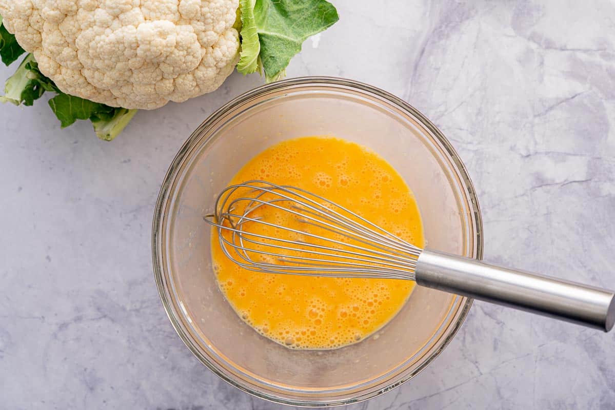 A large glass bowl full of whisked egg sitting on the bench next to a large cauliflower