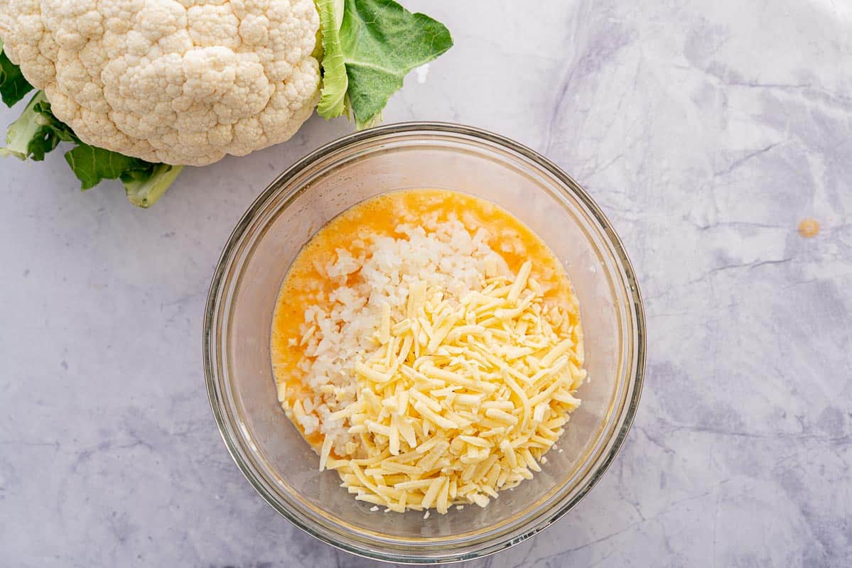 All of the Ingredients in a large glass bowl sitting on a bench next to a cauliflower