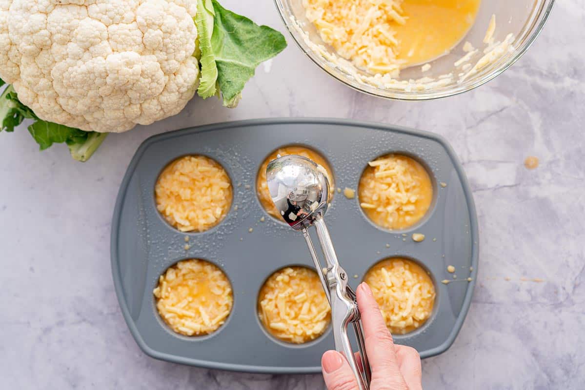 A grey muffin tray filled with scoops of egg muffin mixture on a bench next to a glass bowl of egg mixture.