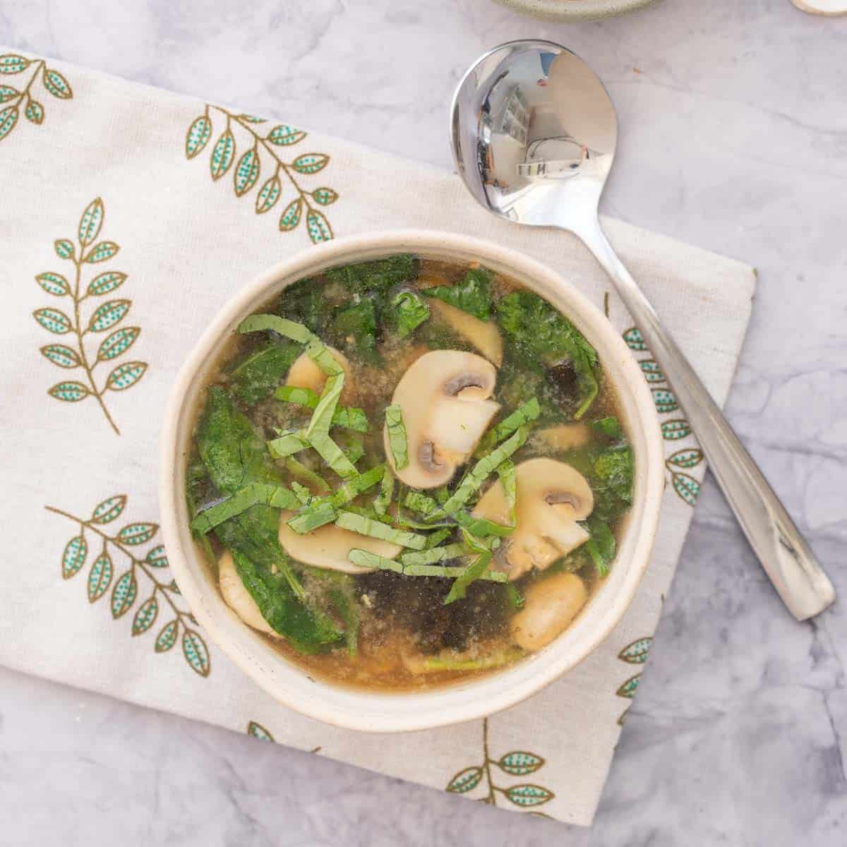 A bowl fulled to the brim with Chicken Miso soo sitting on a tea towel with a spoon next to it.