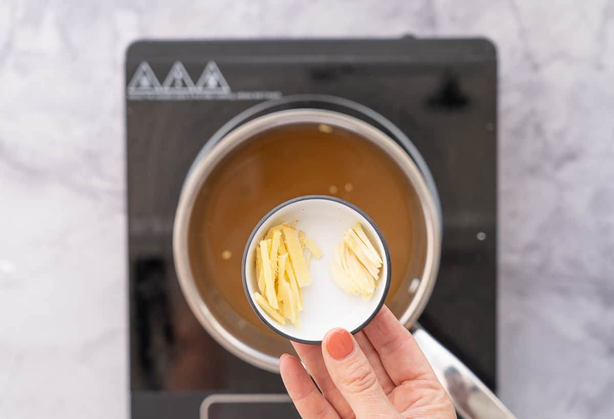 A hand holding a small ramekin of finely sliced ginger above a pot on an element