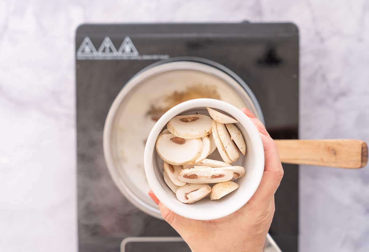 A hand holding a ramekin of sliced mushrooms above a pot on an element