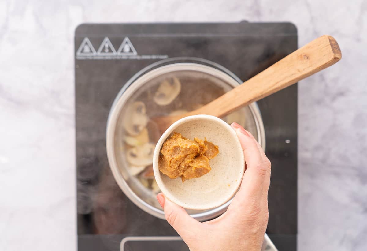A hand holding a small ramekin of miso paste above a pot full of ingredients on the element