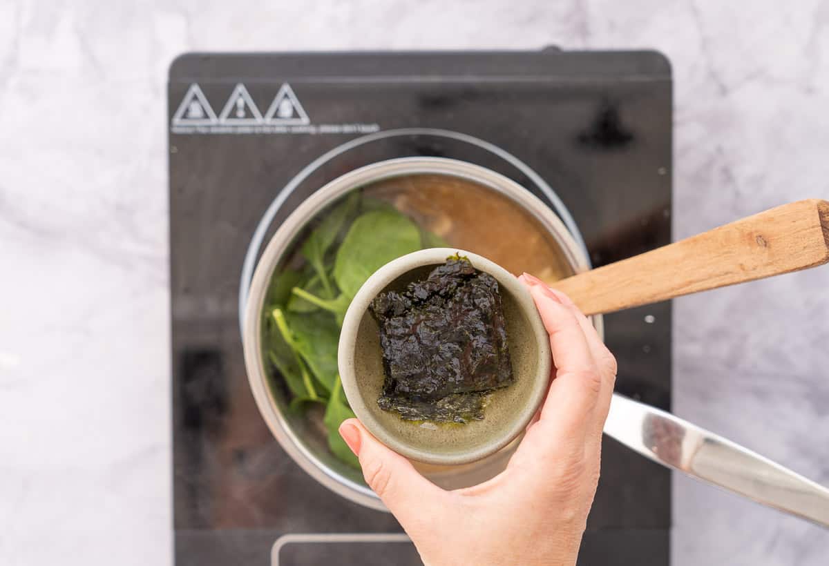 A ramekin of seaweed sheets being held above a pot full of soup on an element