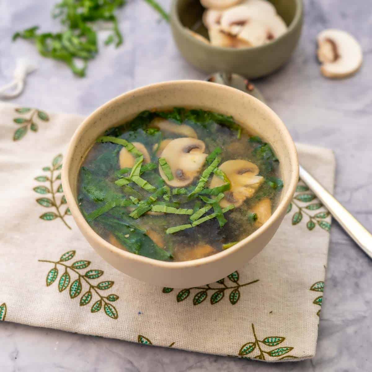 A bowl fulled to the brim with Chicken Miso soo sitting on a tea towel with a spoon next to it. With a bowl of sliced mushrooms in the background