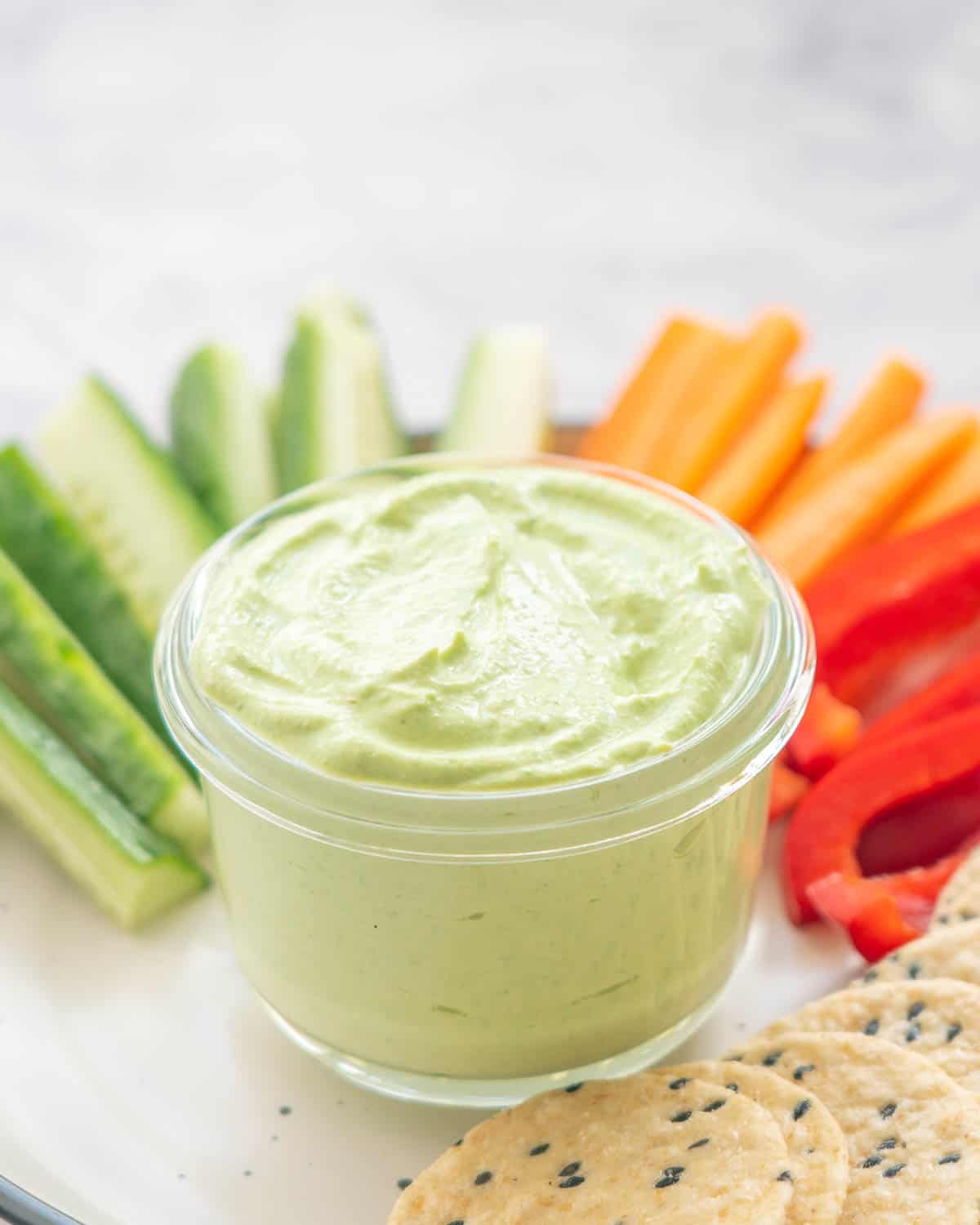 front view of cucumber, carrot and capsicum slices and crackers on a plate with a glass ramekin of cottage cheese dip