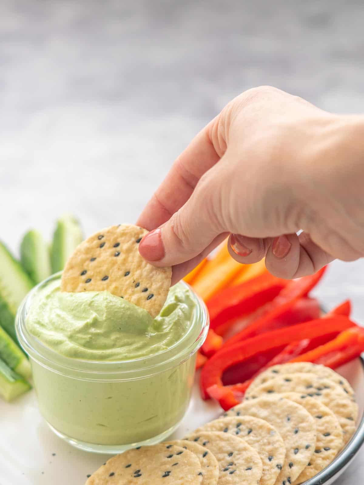 A plate full of crackers and sliced fresh capsicums, carrots and cucumbers with a hand dunking a cracker into a ramekin of Cottage Cheese Dip